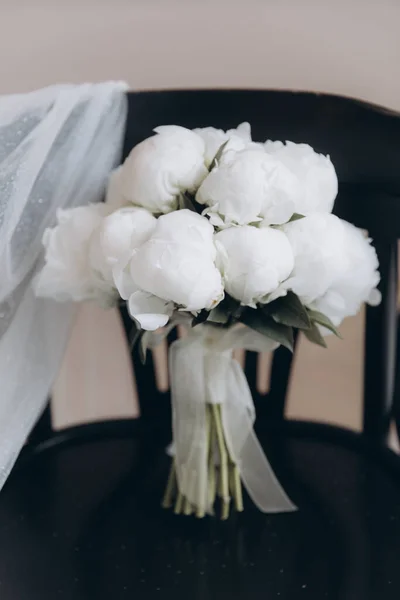 Beautiful white wedding bouquet of peonies on a black chair with a dress and a veil