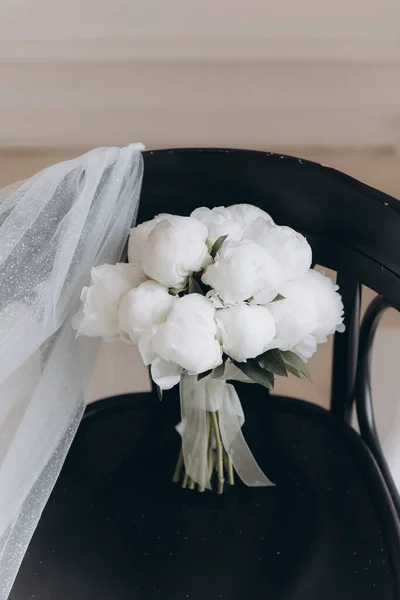 Beautiful white wedding bouquet of peonies on a black chair with a dress and a veil