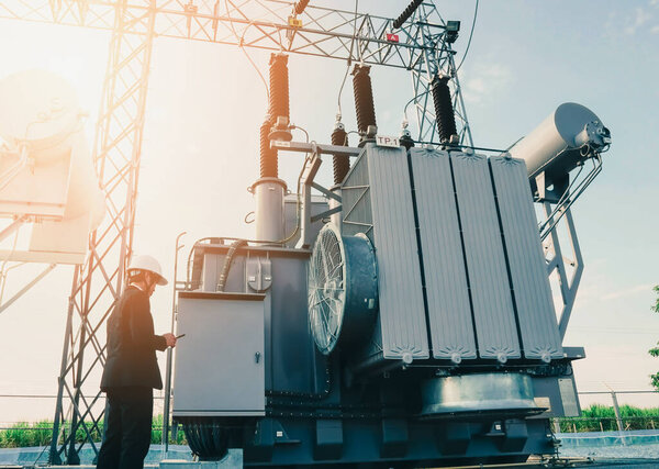 Businessman wearing a black suit, standing looking at a large power transformer with orange sunlight and white sky to be background, Concept about business people who want to invest in energy.