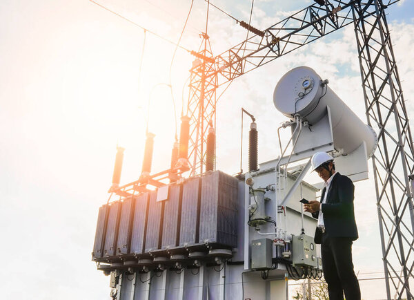 A low angle image of a businessman wearing a black suit, standing looking at a large power transformer with blue sky to be background, Concept about business people who want to invest in energy.