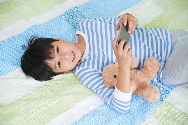 Niño acostado en la cama con teléfono inteligente — Foto de Stock