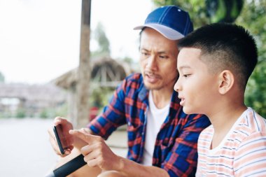 Babam, gölde balık tutarken küçük oğullarına akıllı telefon ekranında balıkların fotoğrafını gösteriyor.
