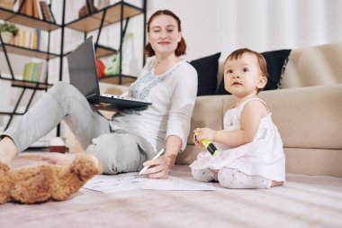 Finansal belgelerle çalışan gülümseyen genç bir kadın ve sevimli küçük bebeğine bakıyor.