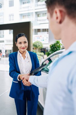 Güzel gülümseyen genç satış müdürü müşteriyle tokalaşıyor, arabaları göstermeden önce onu selamlıyor.
