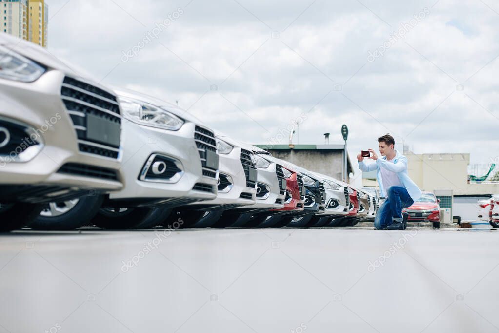 Young man in casual clothes photographing rows of cars in car dealership parking