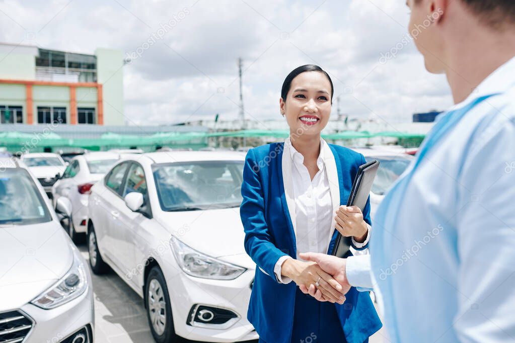 Smiling pretty Vietnamese car dealership saleswoman in suit shaking hand of customer