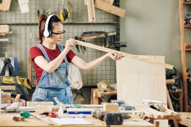 Gözlüklü ve kulaklıklı gülümseyen genç bayan marangoz ahşap ahşabın etkinliğini kontrol ediyor.