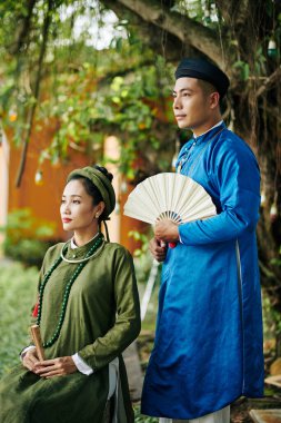 Düşünceli genç Asyalı çift, ellerinde yelpaze, parkta ağacın altında oturuyorlar.