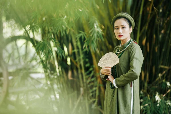 迷人而严肃的年轻女子穿着传统越南服装 头戴木扇 走在户外 — 图库照片