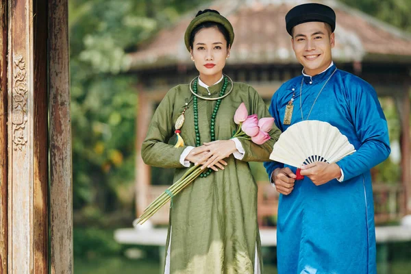 身穿民族服装的越南年轻夫妇站在户外 手中拿着莲花和雕刻的扇子 对着镜头笑着 — 图库照片