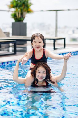 Yüzme havuzunda dururken küçük bir kızı omuzlarında taşıyan neşeli, güzel Asyalı kadın.