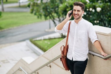 Neşeli, yakışıklı, sakallı bir adam dışarıda dikilirken arkadaşlarıyla telefonda konuşuyor.