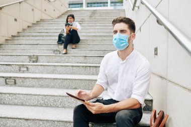 Medikal maskeli düşünceli genç girişimci basamaklarda oturuyor ve dijital tablet üzerinde çalışıyor.