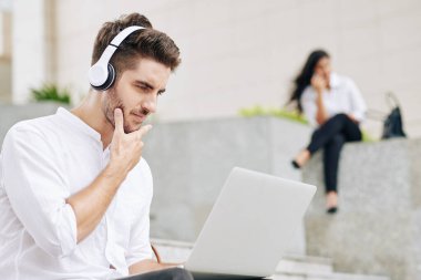 Kulaklıklı genç iş adamının portresi. Rapor ve makaleleri dizüstü bilgisayardan okuyor.