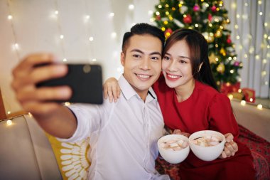 Gülümseyen güzel Vietnamlı çift bir fincan sıcak çikolatayla evde Noel ağacına karşı selfie çekiyor.
