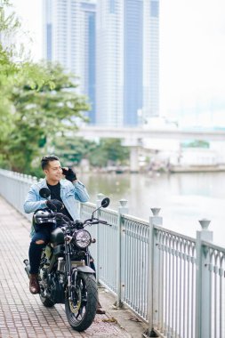 Motosiklette oturan ve telefonda arkadaşlarıyla konuşan mutlu Asyalı adam.