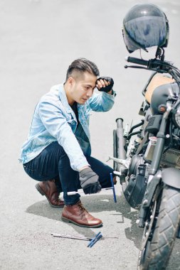 Kafası karışmış genç bir adam bozuk motosiklete bakıyor ve hangi parçanın bozuk olduğunu anlamaya çalışıyor.