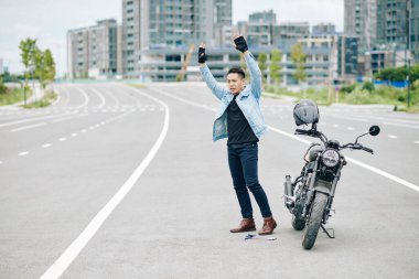 Motosikletli el kaldırıyor ve yolda bozuk motosikletiyle dururken sürücülerin dikkatini çekmeye çalışıyor.