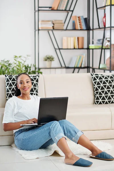 Pretty lovely young Black woman in jeans and t-shirt sitting on the floor with laptop and looking at camera