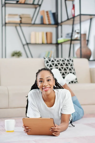 美丽的年轻黑人女人 头发蓬乱地躺在地板上 手里拿着数码平板电脑 对着相机微笑 — 图库照片
