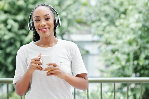 年轻微笑的黑人妇女头戴耳机站在阳台上 端着咖啡 对着相机笑的画像 — 图库照片