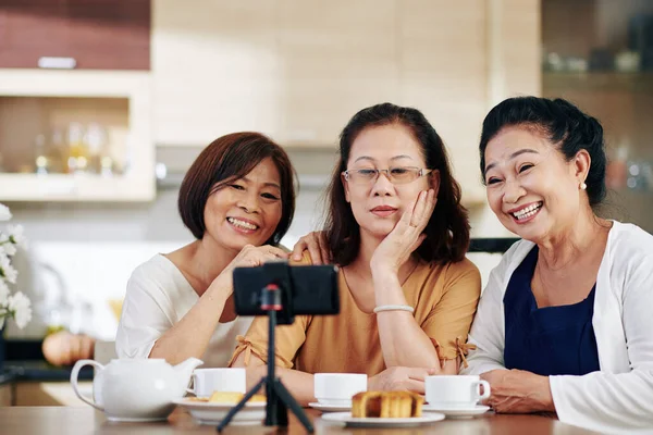 台所のテーブルに座っていると 幸せな高齢者の女性のグループがスマートフォンで自撮り話をする — ストック写真