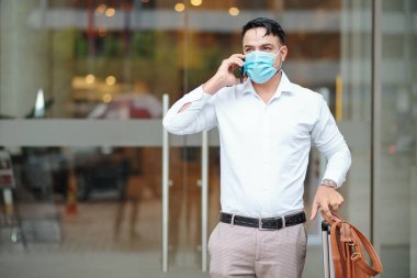 Sağlık maskeli, valizli ve telefonda konuşan ciddi yakışıklı bir adam.