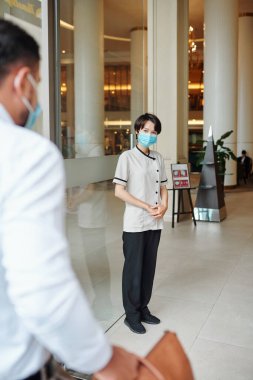 Tıbbi maske takmış gülümseyen otel kapıcısı lobide misafirleri karşılıyor.