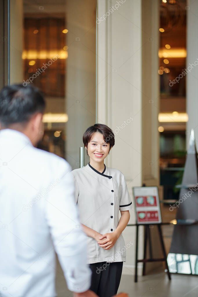 Smiling young Asian concierge looking at man entering hotel lobby