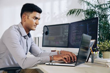 Düşünceli genç Hintli programcı iki monitörle ofis masasında oturuyor ve dizüstü bilgisayarında daktilo yazıyor.