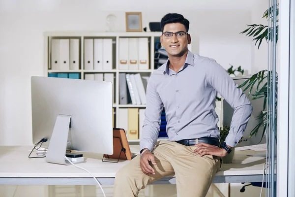 Portrait of young positive Indian businessman sitting on his office table and smiling at camera