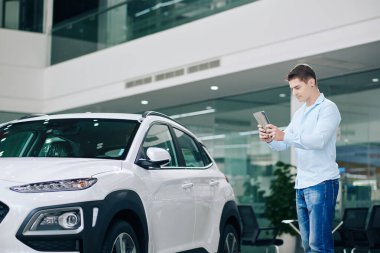 Genç adam araba galerisinde sevdiği arabaların fotoğraflarını çekmek için tablet bilgisayar kullanıyor.