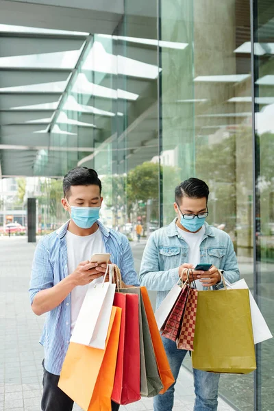 Young Vietnamese men in medical masks walking in the street with shopping bags and texting friends or checking notifications