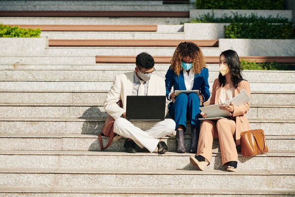 Multi-ethnic business team in medical masks sitting on steps with laptop and documents and working together on project