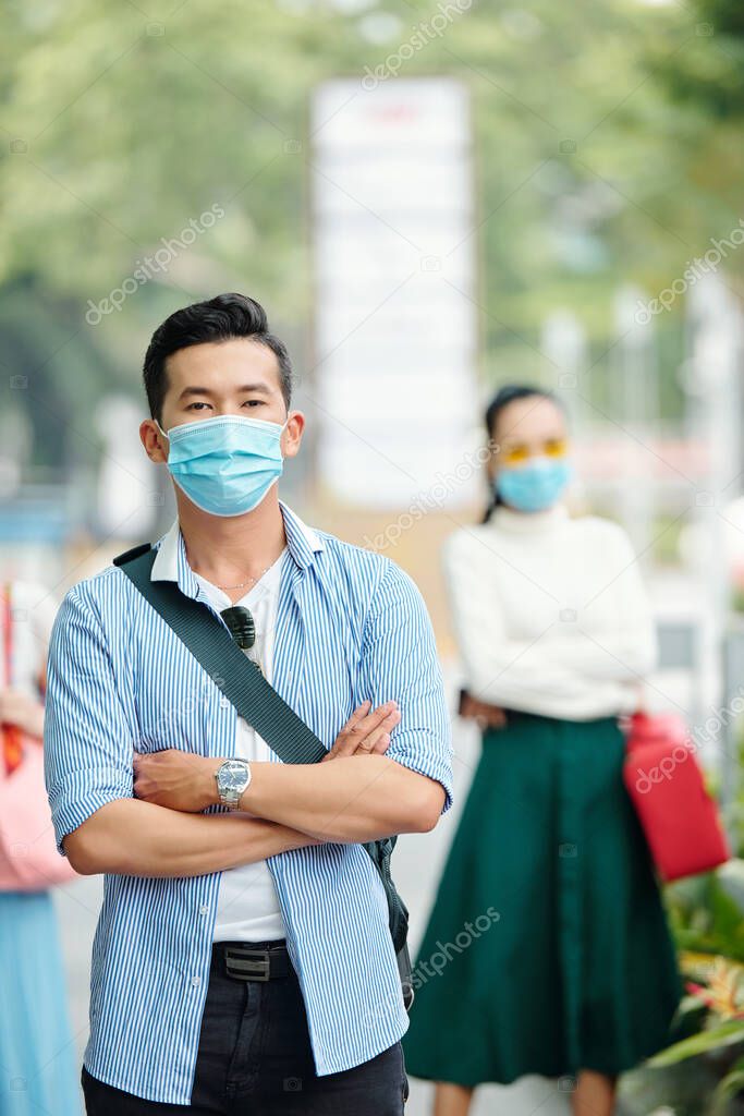Handsome confident young Vietnamese man in medical mask standing outdoors with arms folded and looking at camera