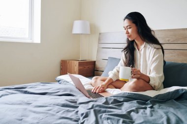 Salgın yüzünden evden çalışan güzel bir genç kadın, yatakta oturup kahve içiyor ve e-postalarını kontrol ediyor.