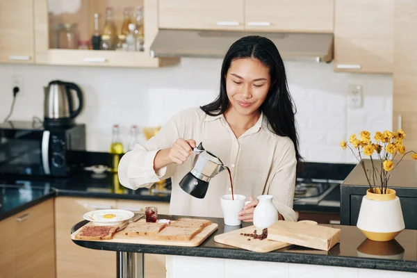 在厨房吃早餐的时候 笑着把咖啡倒在杯子里的漂亮中国女人 — 图库照片