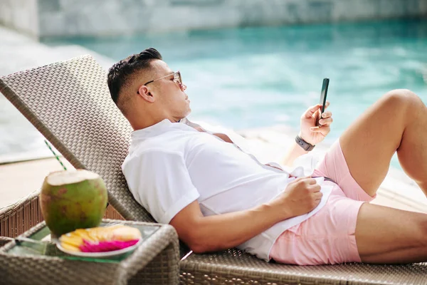 スイミングプールでチェースラウンジで休んでいる男とテキストメッセージを読むか スマートフォンで通知をチェック — ストック写真