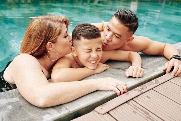Parents kissing their happy laughing preteen son on both cheeks when standing in swimming pool
