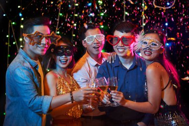 Bir grup neşeli Asyalı komik karnaval maskeli genç konfeti altında durup şarap kadehleriyle kadeh kaldırıyor.