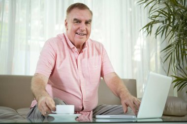 Gülümseyen son sınıf öğrencisinin portresi. Bir fincan kahve içip evde dizüstü bilgisayarla çalışıyor.