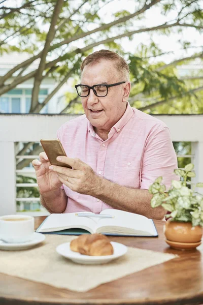 戴眼镜的老人坐在咖啡桌旁 开着笔记本电脑 用智能手机查看留言 — 图库照片