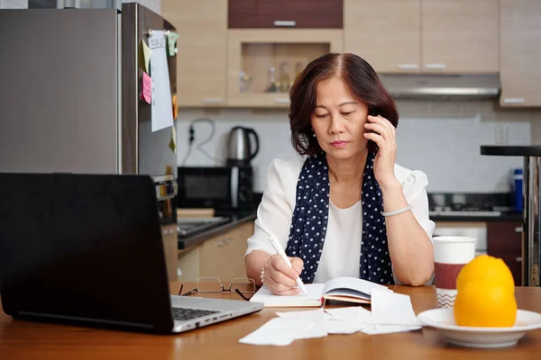Seriöse Seniorin Sitzt Küchentisch Telefoniert Und Macht Sich Notizen — Stockfoto