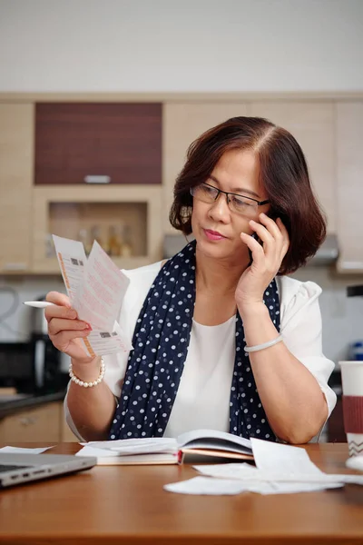 严肃的老年妇女坐在厨房餐桌前打电话和检查公用设施账单的肖像 — 图库照片