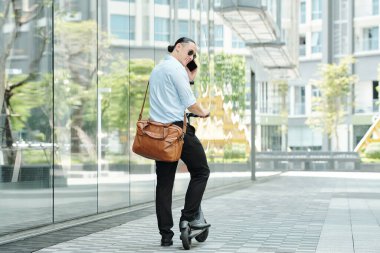 Güneş gözlüklü yakışıklı genç adam elektrikli scooter kullanıyor ve telefonla arıyor.