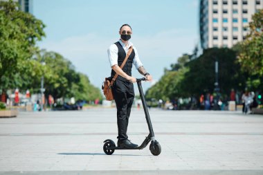 Tıbbi maskeli şık yakışıklı işadamı şehirde elektrikli scooter sürmenin tadını çıkarıyor.