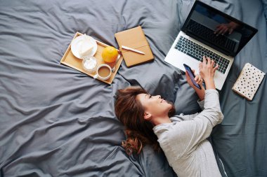 Güzel, gülümseyen, mesajları okuyan ve yatakta kahvaltı ederken bilgisayardaki e-postaları cevaplayan genç bir kadın.