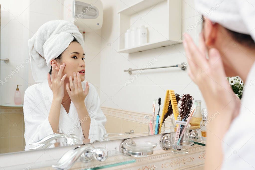 Young Asian woman standing at bathroom mirror and looking at blemishes on her skin after taking morning shower