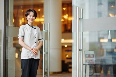 Güzel gülümseyen kadın otel odacısının portresi cam kapılarda dikilip kameraya bakıyor.