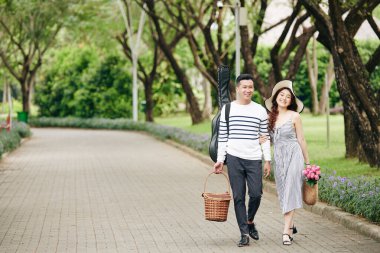 Piknik sepetleri ve gitarlarıyla şehir parkında yürüyen mutlu bir Asyalı çift.